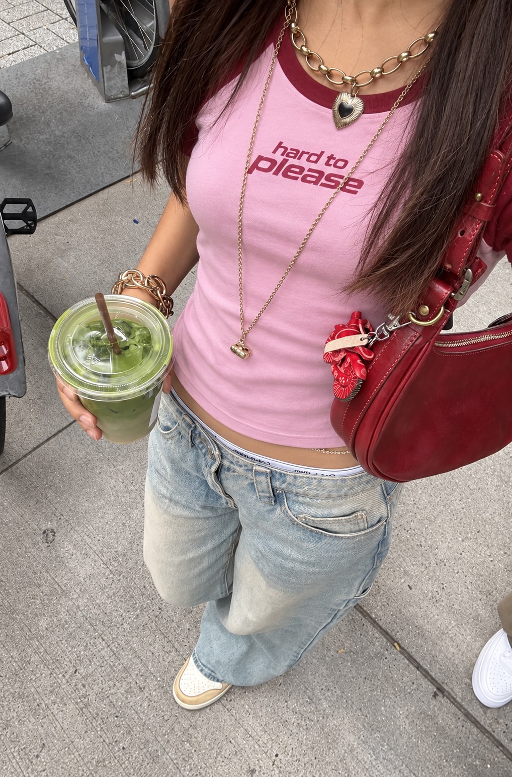 Hard to Please pink and burgundy baby tee worn on a sidewalk, shot from above with a matcha drink and a cherry red bag