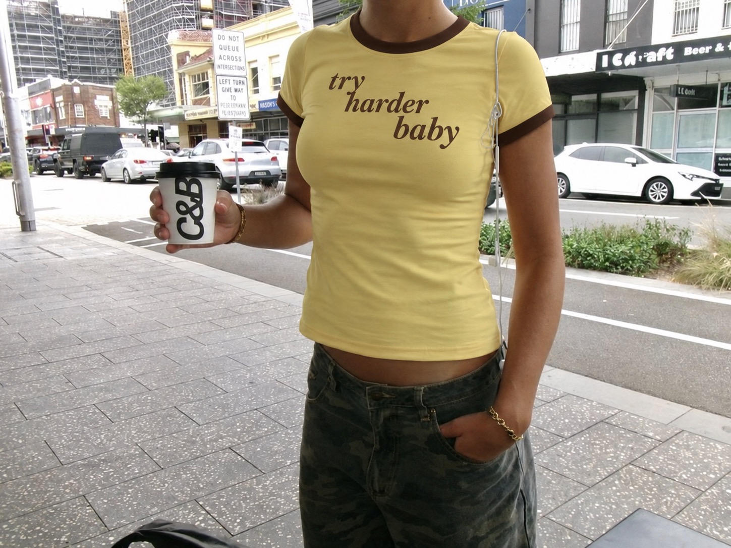 Try Harder Baby yellow and brown baby tee worn on a city street, holding a takeaway coffee